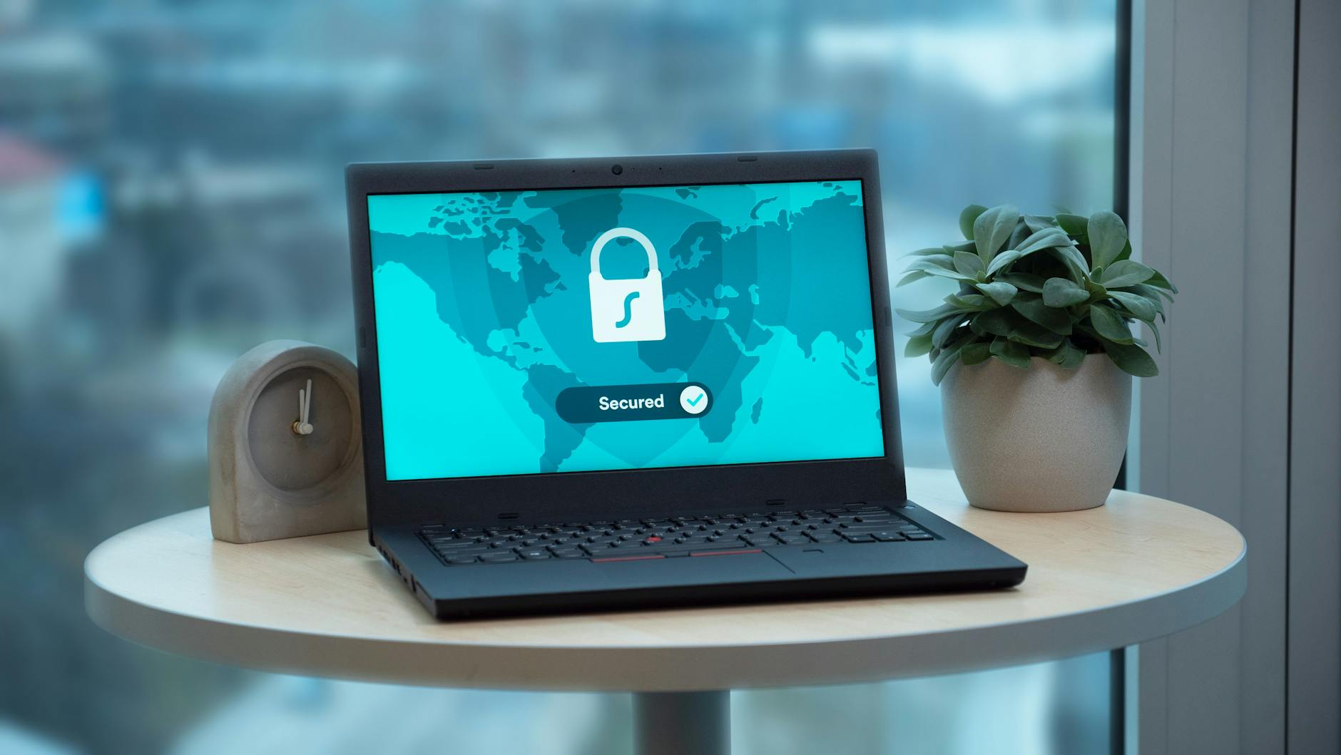 Laptop z ikoną kłódki na biurku obok rośliny i zegara